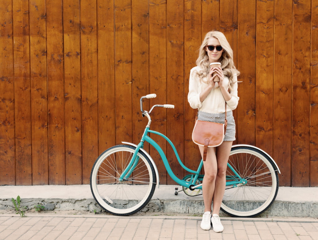 Young sexy blonde girl with long hair with brown vintage bag in sunglasses standing near vintage green bicycle and holding a cup of coffee, have fun and good mood looking in camera and smiling